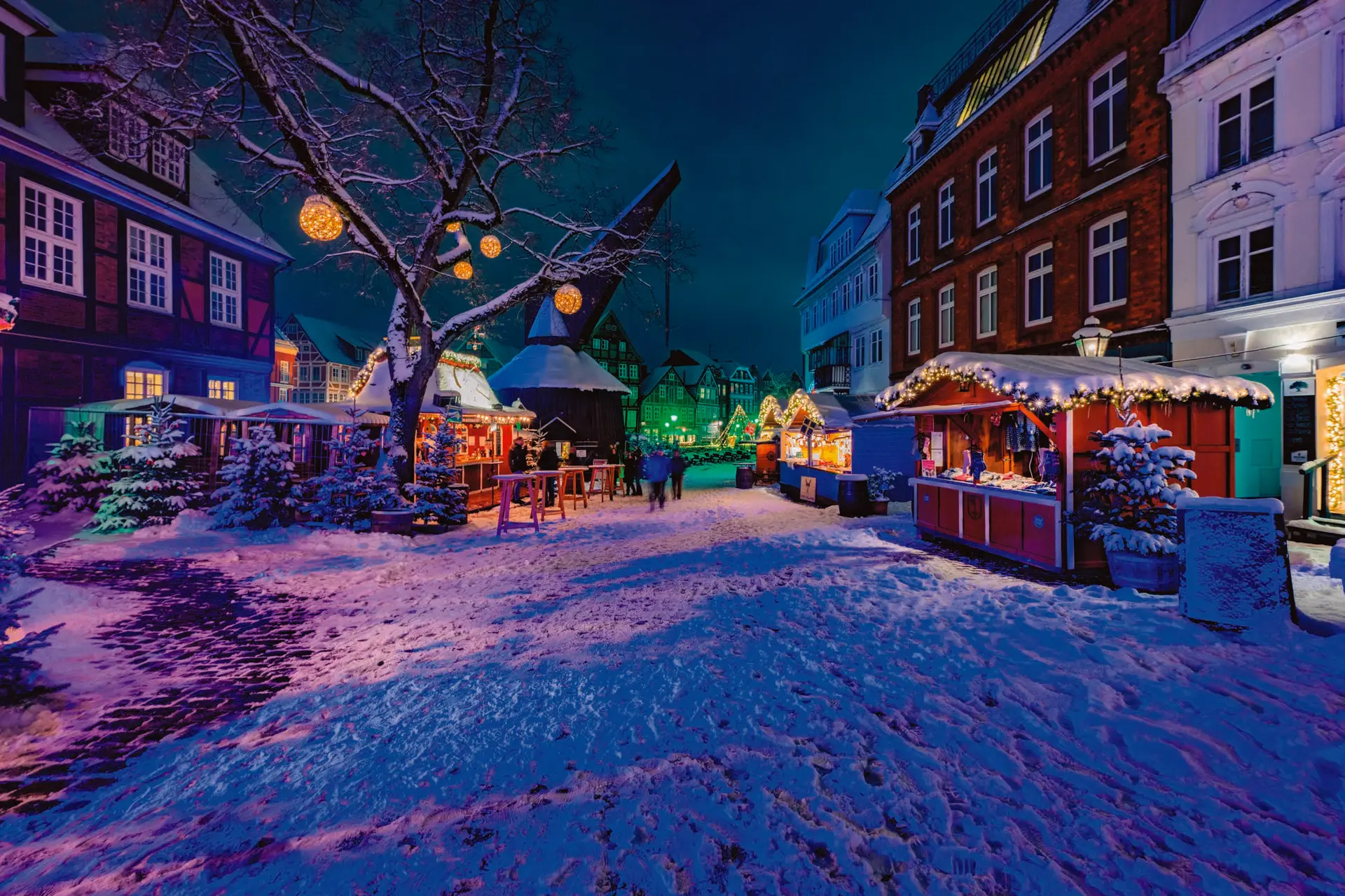 Der verschneite Stader Fischmarkt bei Nacht: Weihnachtsbuden, der alte Holzkran und ein Baum mit leuchtenden Lichterkugeln